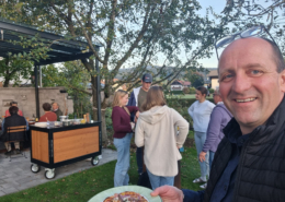 Ein lächelnder Mann hält einen Teller mit Pizza in die Kamera, während sich im Garten mehrere Besucher unterhalten.