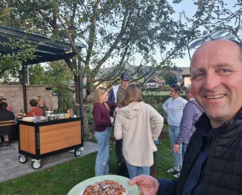 Ein lächelnder Mann hält einen Teller mit Pizza in die Kamera, während sich im Garten mehrere Besucher unterhalten.